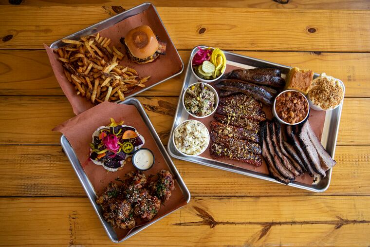 Pictured are the smash burger topped with a house pork belly and a side of fries, two vegetable sides of smoked beats and tempura fried cauliflower, and a platter of smoked meats including sausage, ribs, and brisket, with a side of mac and cheese, potato salad, smoked pit beans, pickles, corn bread, and slaw at Wildwoods BBQ.