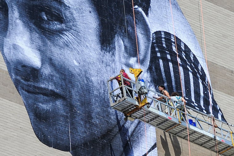 Artist JR, sporting his fedora and sunglasses, installs his mural of Ibrahim on the side of the Graham building at 30 S. 15th St. in Philadelphia on Thursday, July 23, 2015. (MICHAEL PRONZATO / Staff Photographer)