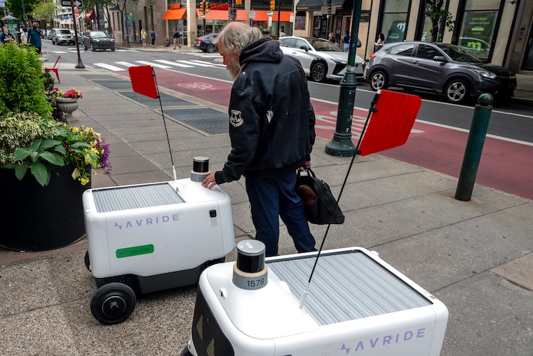 After asking if it has a name, pedestrian Gary Wallace “pets” an Uber delivery robot (left) joined by a second one, parked and apparently waiting for an order in the 1900 block of Chestnut Street.