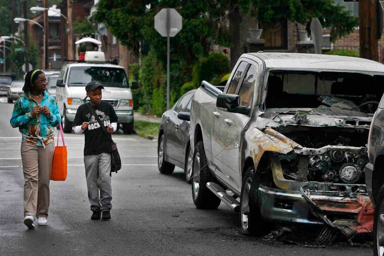 Pedestrians pass one of seven vehicles that were set ablaze early Wednesday on Georges Lane and West Berks Street in Wynnefield. A resident said an explosion woke her at 4:30 a.m.
