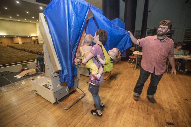 Vera Dugan, 18 months, right, holds on tight to Bridget Huffman, center, as she holds her son Drew Huffman, 20 months, left, as they all go into the voting booth together, on the stage of the auditorium of South Philadelphia High School on May 15, 2018. Nazim Karacaa, right, holds open the blue curtain so Huffman and her brood can enter to vote in the primary election in the 39th District in South Philadelphia.