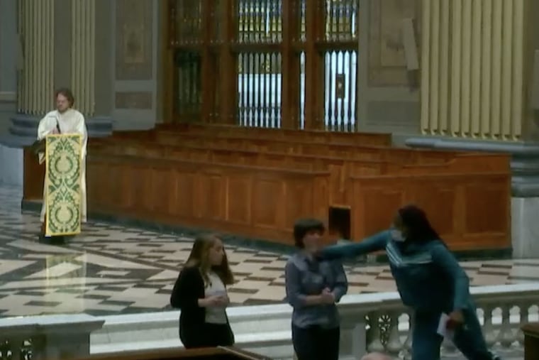 In this screenshot from the livestream of Sunday's Mass at the Cathedral Basilica of SS. Peter and Paul, a woman in a green track suit is shown punching a lector during the service. Philadelphia police said Monday that they had identified the suspect but had declined to pursue charges against her.