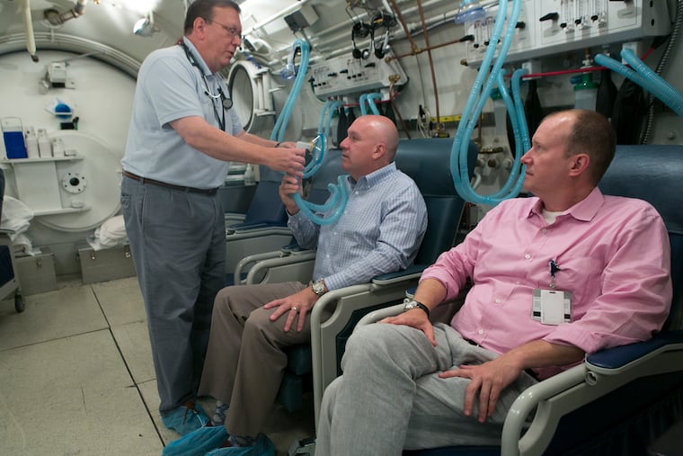 Inside Penn's submarine-like hyperbaric medicine chamber, Dr. Kevin Hardy (left) holds an an oxygen mask in front on former patient David Wolfe, as Dr. Matt Kelly looks on.