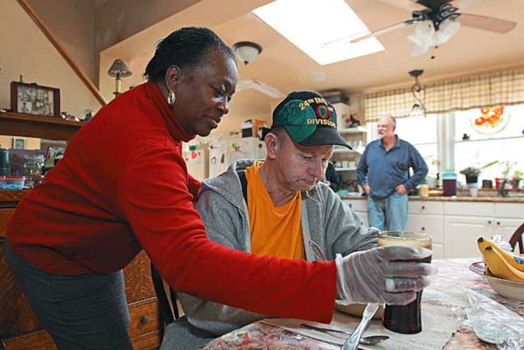 Maedell Cuffy, left, a residential aide, brings Carl Ferrell, center, his drink as he sits down to lunch at the family table in the Borger home. This is the home of Ron Boger who is a medical foster home for vets provider. A new U.S. Department of Veterans Affairs program offers aging and infirmed vets who have no family to care for them the chance to live in a private home and avoid a nursing facility. The Medical Foster Home care program places vets with families who provide room, board and personal care. The families are paid for their services. VA hospitals in Coatesville and Philadelphia offer the service. PVETS27 01/23/2013 ( MICHAEL BRYANT / Staff Photographer )