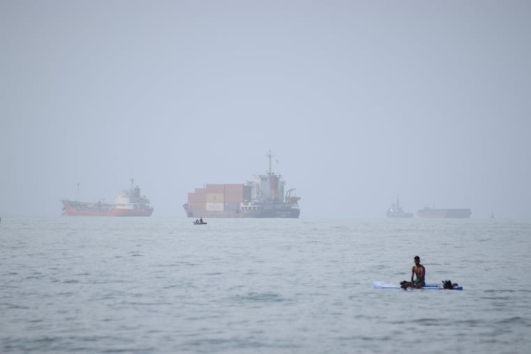 A man sits on a surfboard as a container ship and other commercial vessels appear anchored in the Strait of Hormuz off Bandar Abbas, Iran, Monday, July 27, 2026.