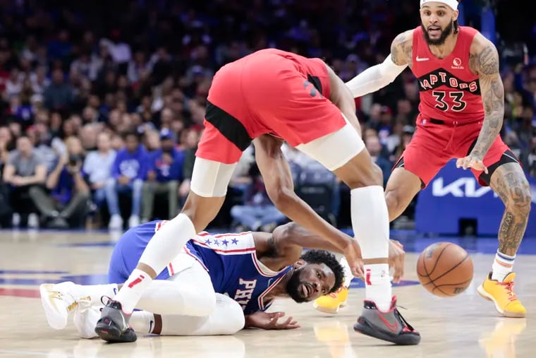 Sixers center Joel Embiid reaches for the loose basketball on the floor against Toronto Raptors forward Scottie Barnes and Toronto Raptors guard Gary Trent Jr.