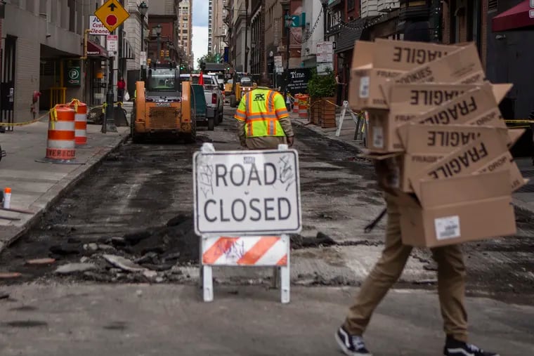 Pedestrians walk by the closed-off intersection at 13th and Sansom Streets, nearly a year after last summer’s 48-inch water main break.